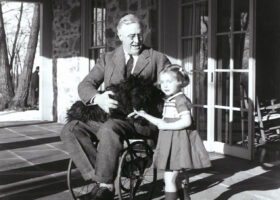 Black and white image of President Franklin Roosevelt in a wheelchair with a dog on his lap and young girl standing next to him.