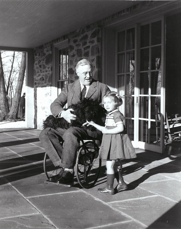 Black and white image of President Franklin Roosevelt in a wheelchair with a dog on his lap and young girl standing next to him.